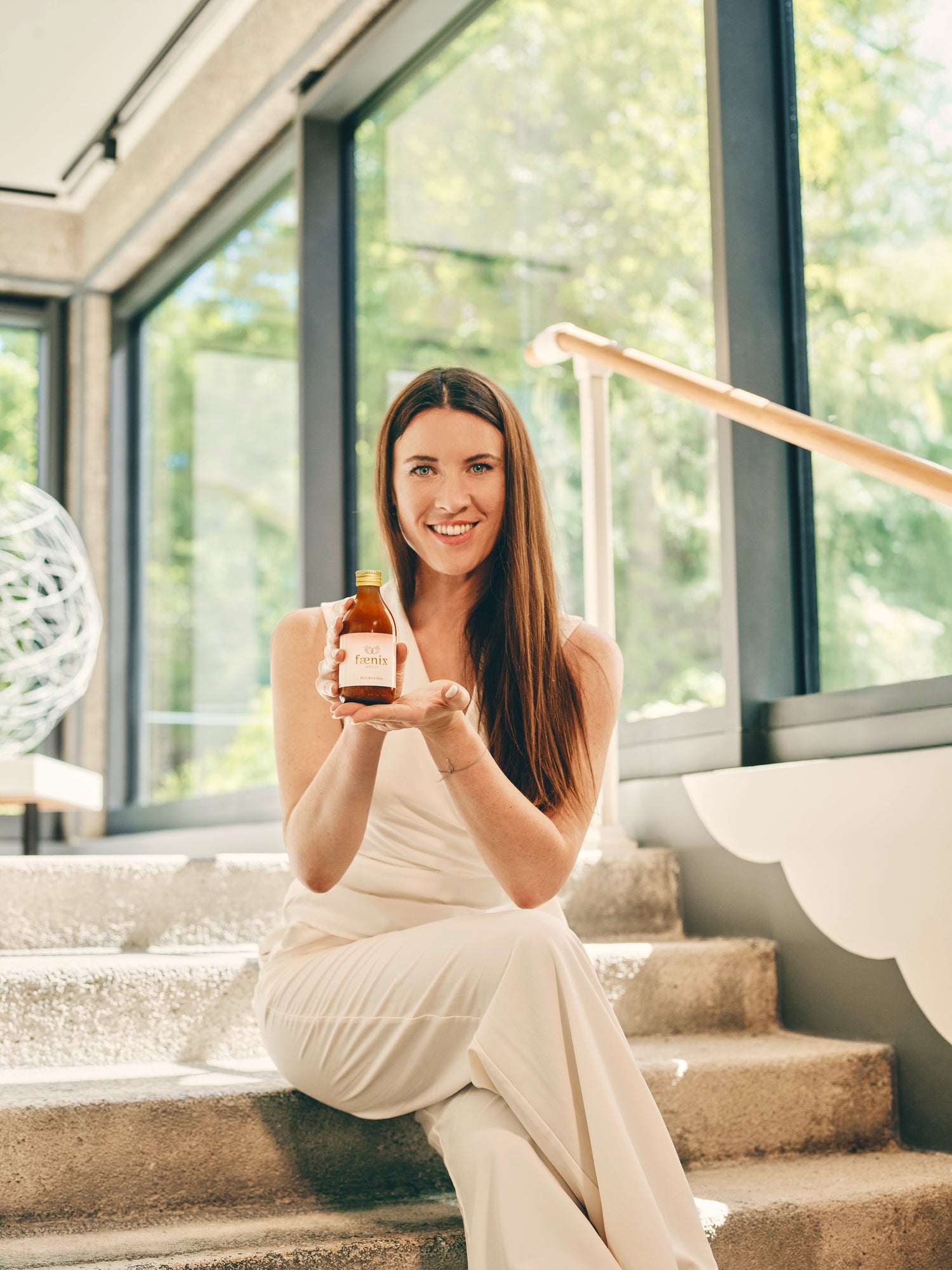 Woman in a white dress holding a bottle in a modern indoor setting with large windows.