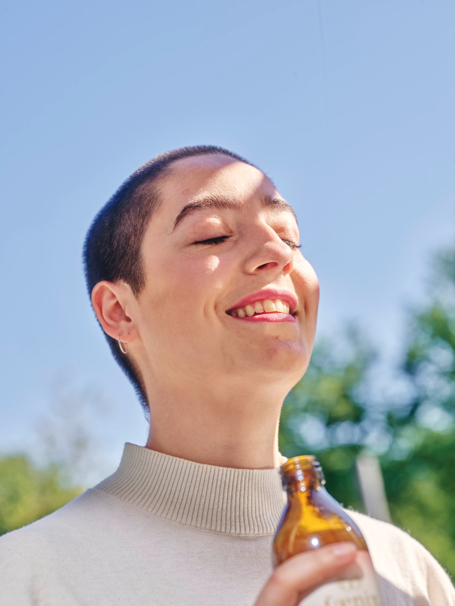 Person holding a brown bottle against a blue sky and greenery background
