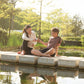 Two people sitting on a dock by a pond with trees in the background