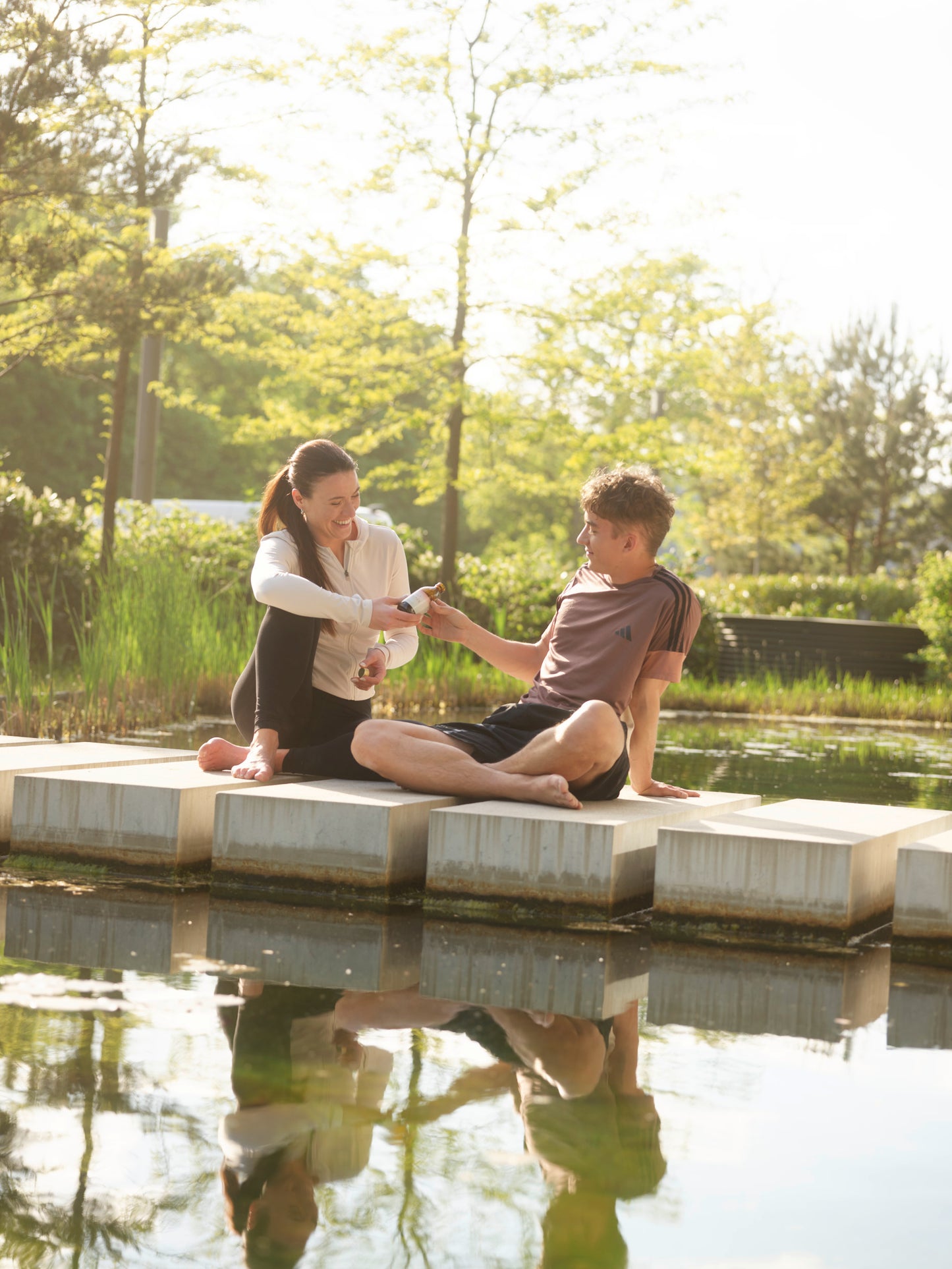 Two people sitting on a dock by a pond with trees in the background