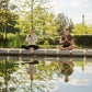 Two people meditating by a pond with trees in the background