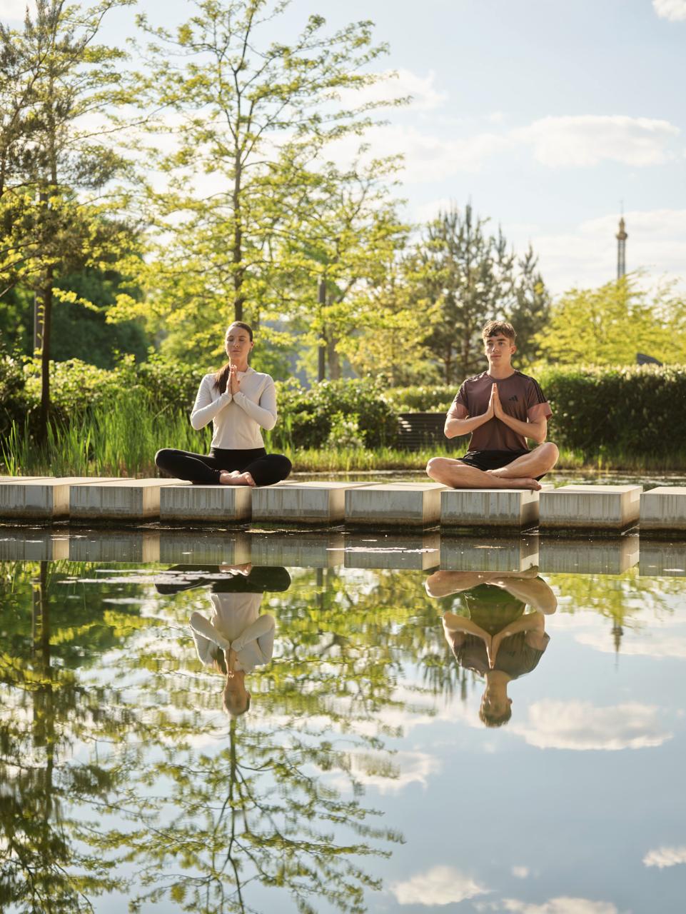 Two people meditating by a pond with trees in the background