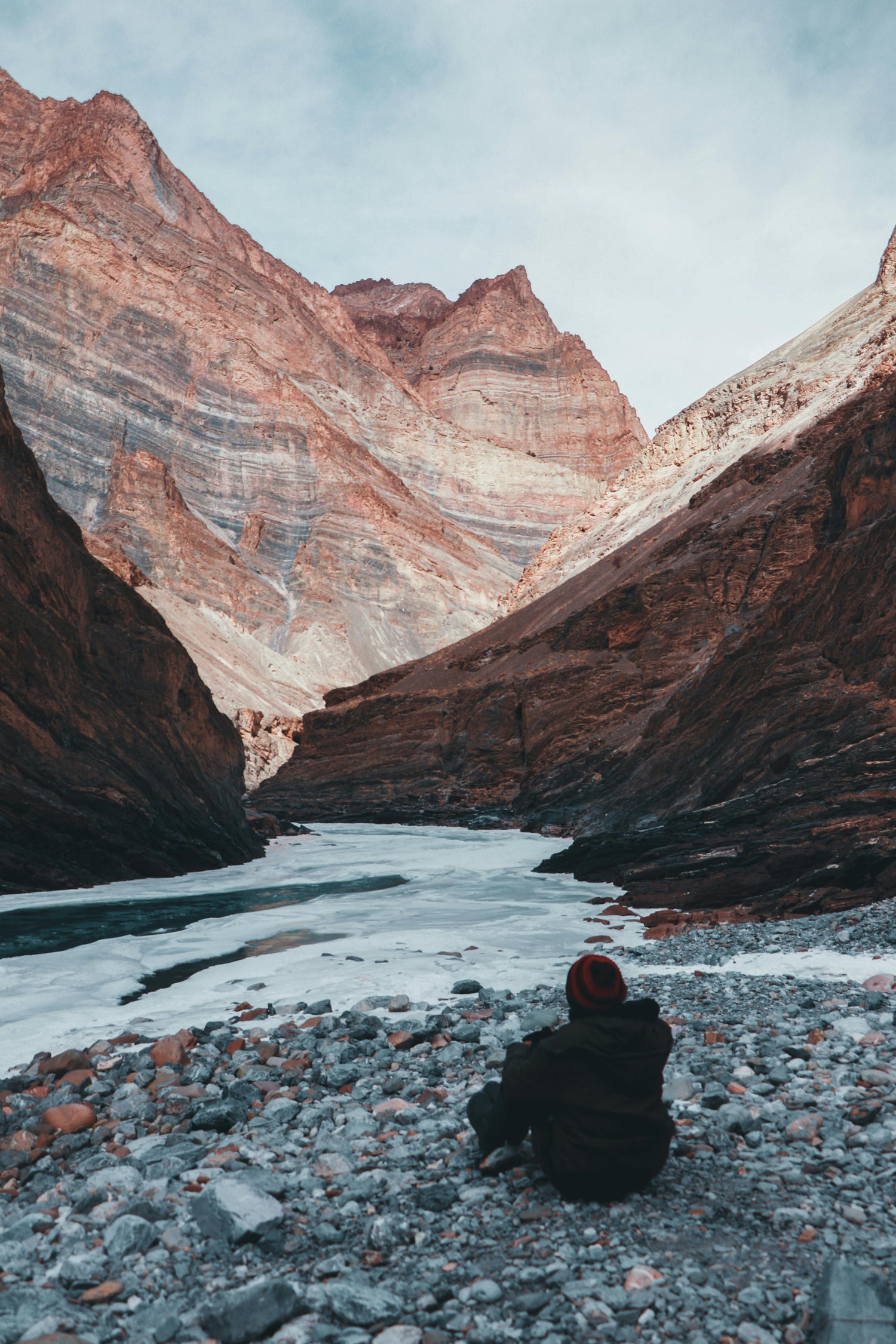 Person sitting on a rocky outcrop in a vast canyon with a river and mountains.