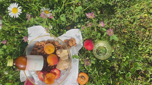 apricots, almonds and a bottle in a bow on a law, someone is pouring faenix elixir in an empty glass